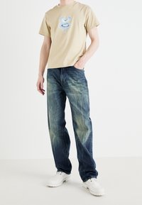 Camiseta de algodón beige con gráfico frontal, combinada con vaqueros desgastados azul claro. Modelo lleva zapatos blancos. Diseño minimalista y casual.