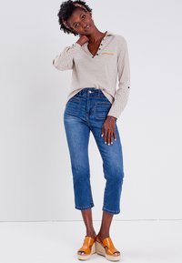 Blusa de lunares beige claro con cuello y botones, combinada con jeans de tiro alto azules. La modelo lleva sandalias de plataforma naranjas.