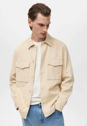 Beige button-up shirt with two front pockets, made of lightweight fabric, featuring a relaxed fit and a pointed collar. Worn over a white t-shirt.