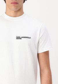 Weißes Baumwoll-T-Shirt mit Rundhalsausschnitt und kurzen Ärmeln. Schwarzes Logo "KARL LAGERFELD JEANS" auf der linken Brust. Weiche Textur.