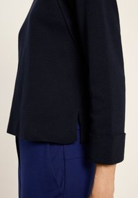 Seitenansicht einer Person, die einen dunkelblauen Pullover mit gekrempelten Ärmeln und blauen Hosen vor einem neutralen Hintergrund trägt.