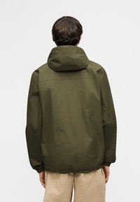 Άνδρας με κοντά σκούρα μαλλιά που φοράει ελιτί πράσινο μπουφάν με κουκούλα και μπεζ σορτς, στέκεται με την πλάτη προς τα εμπρός σε απλό φόντο.