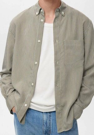 Mężczyzna ubrany w jasnobrązową i białą koszulę w pionowe paski zapinaną na guziki, na białej prążkowanej koszulce na ramiączkach, niebieskie dżinsy, dłonie w kieszeniach.