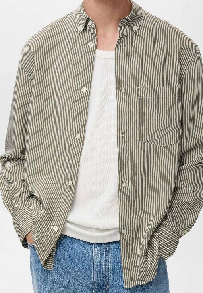 Homme portant une chemise à rayures verticales marron clair et blanches boutonnée, par-dessus un débardeur blanc côtelé avec un jean bleu, mains dans les poches.