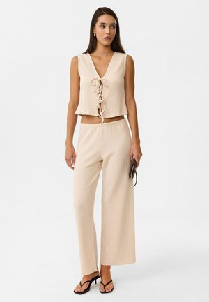 Tweedelig outfit in lichtbeige. De top heeft een V-hals met een voorknoop, en de losse broek heeft een rekbare tailleband. Textuurstof.