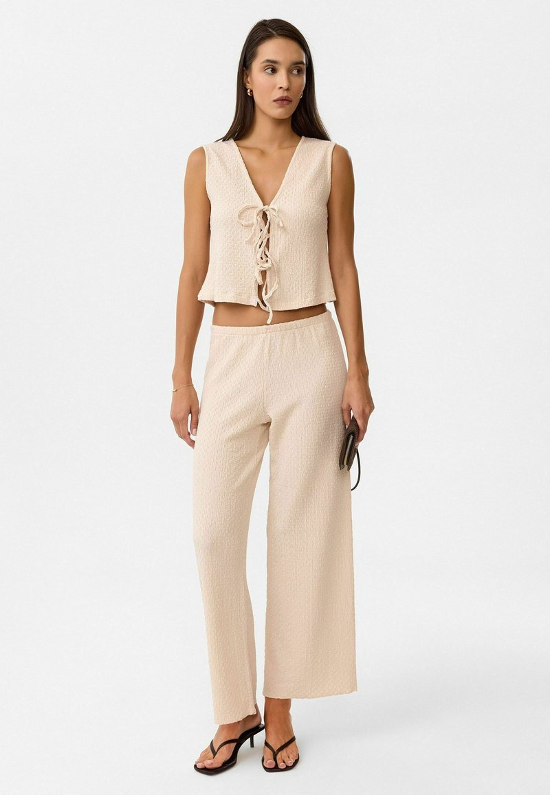 Zweiteiliges Outfit in hellem Beige. Das Oberteil hat einen V-Ausschnitt mit einer vorderen Krawatte, und die locker sitzende Hose hat einen dehnbaren Bund. Strukturiertes Material.