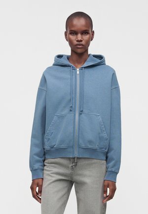 Person trägt einen blauen Kapuzenpullover mit Reißverschluss und hellgraue Jeans und steht vor einem schlichten hellen Hintergrund.