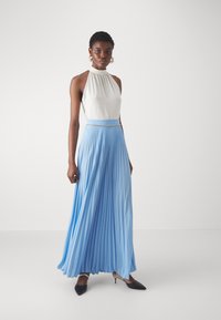Haut à col halter blanc associé à une jupe plissée longueur cheville en bleu clair. La jupe présente un éclat subtil et un accent en chaîne argentée à la taille.