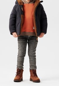 Niebieska kurtka z podszewką z brązowego sztucznego futra, pomarańczowa bluza z kapturem, szare dżinsy i brązowe buty nawiązujące do stylu lace-up. Teksturowane materiały i wyraziste kolory.