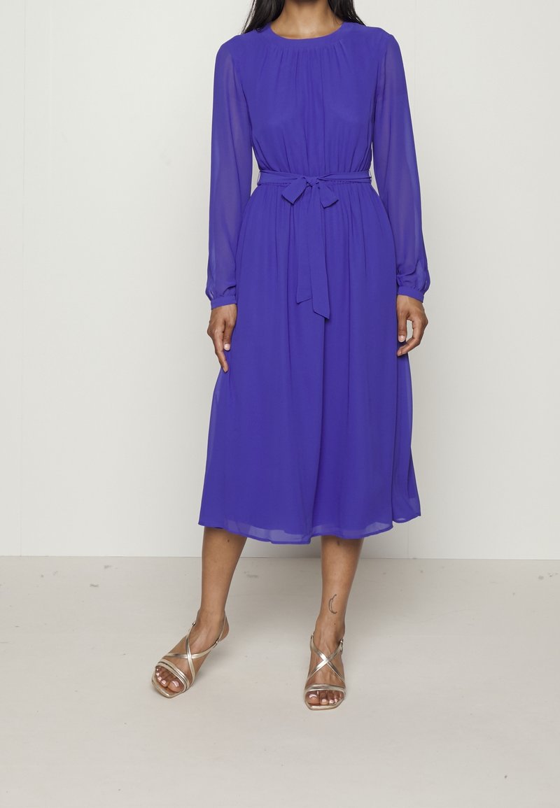 Femme portant une robe violette à manches longues, longueur genoux, avec une ceinture nouée à la taille et des sandales dorées à brides, debout devant un fond uni.