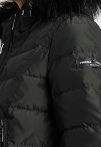 Svart puffjacka med kviltad textur, med en huva i fuskpäls och dragkedja. Märkesetikett på vänster ärm.