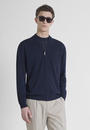Marine blauw ritsvest met een ronde kraag, geribde manchetten en zoom. Heeft een subtiele logo op de borst en een soepele textuur. Model draagt een zonnebril.