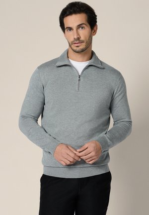 Mężczyzna o ciemnych włosach, ubrany w jasnoszary sweter z półzamkiem na półtorej długości, pod którym ma białą koszulkę, oraz czarne spodnie, stoi na beżowym tle.