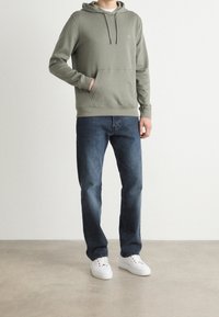 Sudadera con capucha en algodón verde apagado con bolsillo frontal y cordones ajustables, combinada con vaqueros oscuros y zapatillas blancas.