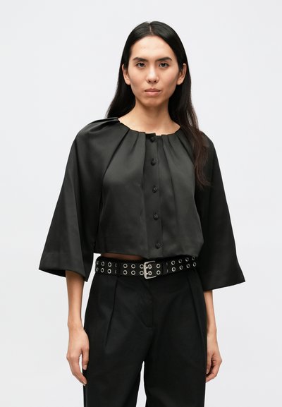Blouse noire courte avec un col rond, des détails plissés et des boutons noirs, associée à un pantalon noir taille haute et une ceinture cloutée.