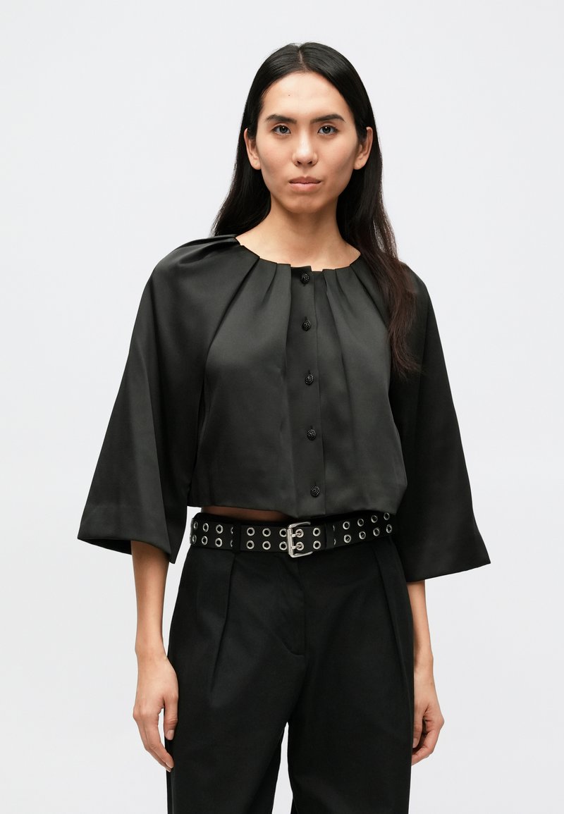 Schwarze kurz geschnittene Bluse mit rundem Ausschnitt, Falten-Details und schwarzen Knöpfen, kombiniert mit hoch taillierten schwarzen Hosen und einem mit Nieten besetzten Gürtel.