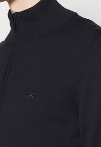 Svart, höghalset pullovertröja i texturerad stickad kvalitet; har en halvdragkedja och en subtil broderad logotyp på bröstet.