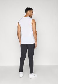 Witte mouwloze tanktop gecombineerd met donkergrijze broek. Model staat met de rug naar de camera, waardoor het ontwerp aan de achterkant goed zichtbaar is. Witte sneakers maken de outfit compleet.