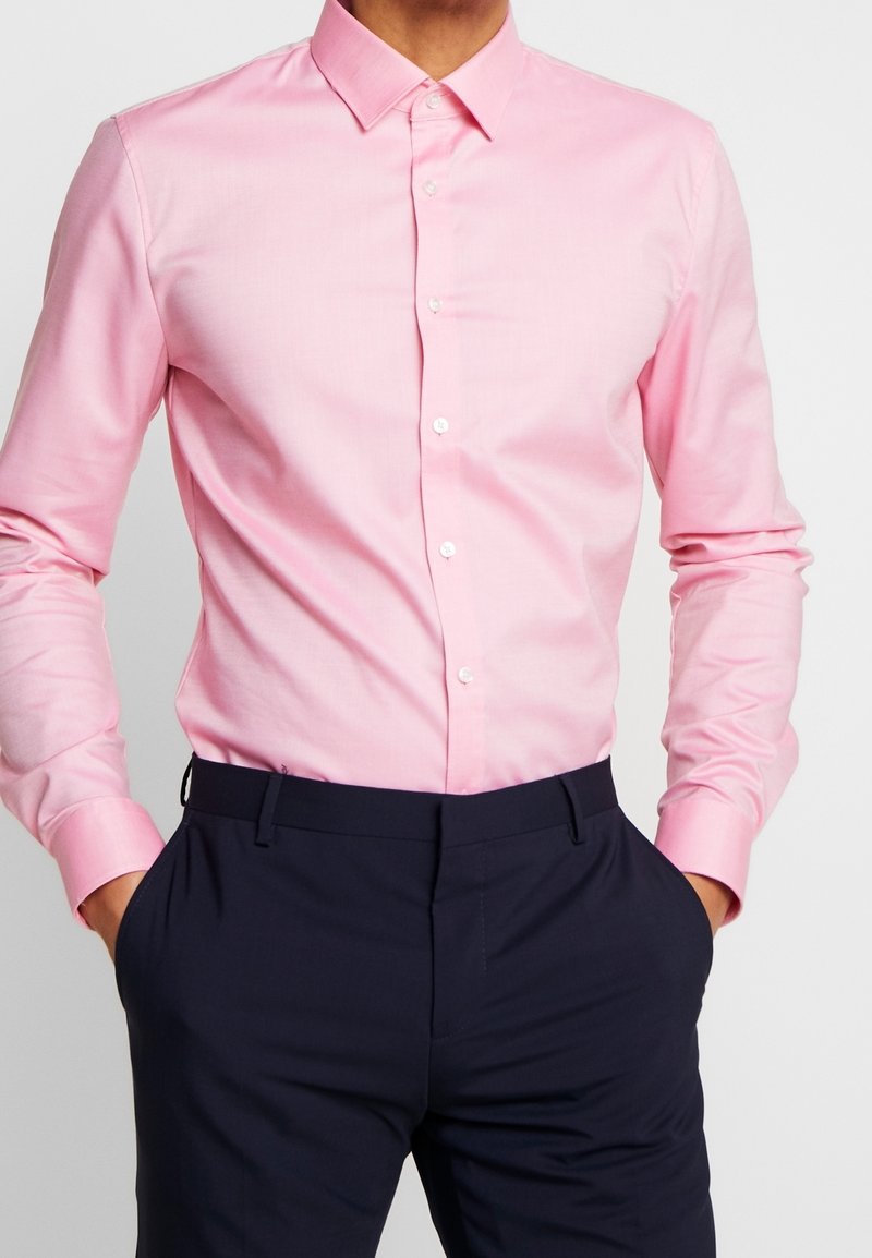 Chemise rose pâle à boutons avec col classique et manches longues, assortie à un pantalon bleu marine foncé, présentant une texture de tissu lisse.