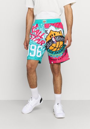 Mann trägt bunte NBA All-Star 1996 San Antonio Shorts mit Basketball-Grafik, weiße Socken und weiße Turnschuhe, steht vor einem einfarbigen Hintergrund.