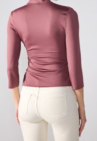 Mauve-värinen napakka paita, jossa on kolmivartiset hihat ja korkea kaulus, yhdistettynä valkoisiin slim fit -farkkuihin, esittäen sulavia pintarakenteita ja minimaalista yksityiskohtaisuutta.