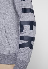 Grå sweatshirt med en texturerad tyg. Har en stor mörkblå tryckt text på den högra ärmen. Inkluderar en känguruficka.