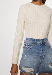 Pullover in maglia a treccia di colore crema con maniche lunghe abbinato a pantaloni shorts in denim a vita alta, con effetti graffiati e orli sfrangiati, di un lavaggio azzurro chiaro.