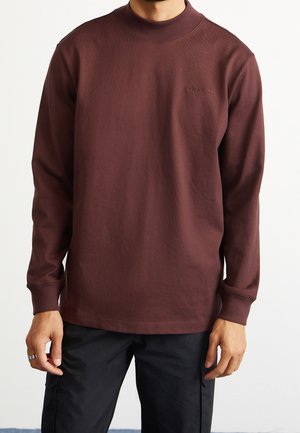 Man draagt een bordeauxrode Adidas sweater met lange mouwen en een zwarte broek, staand tegen een effen witte achtergrond.