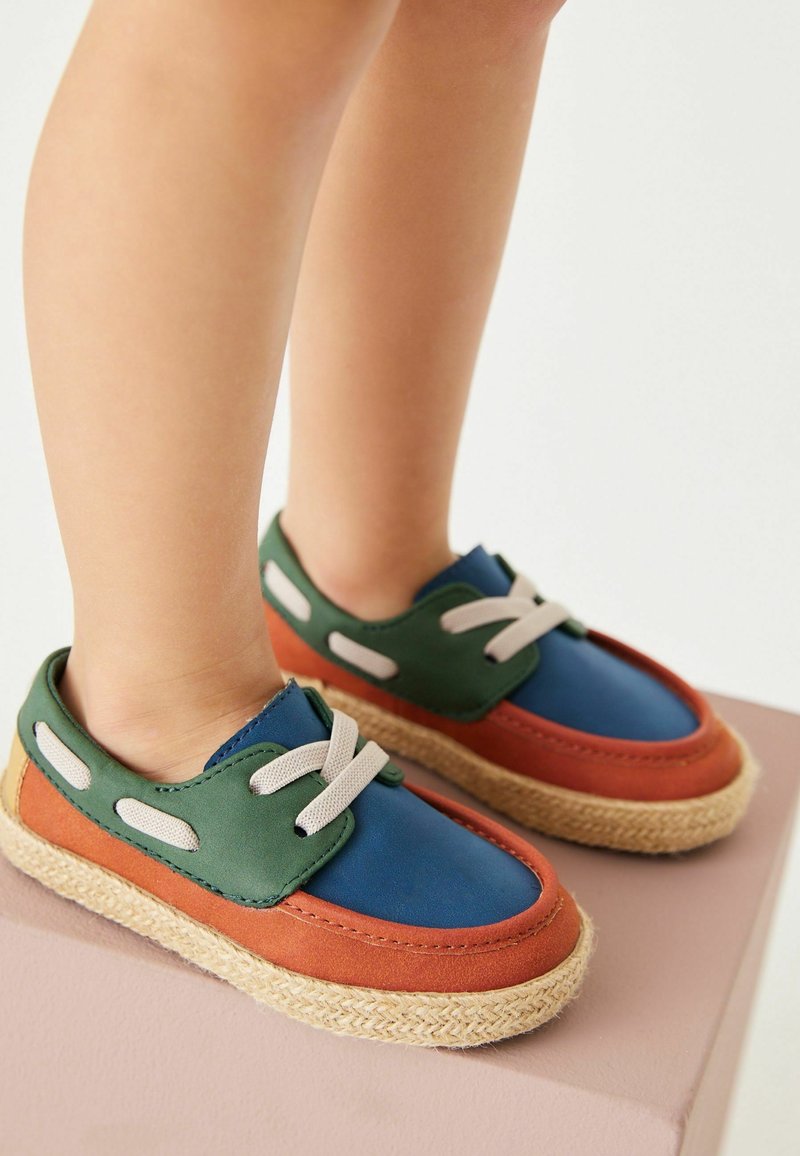 Kolorowe buty z niebieską i zieloną cholewką, pomarańczowymi akcentami oraz teksturowaną podeszwą z juty. Sznurówki są jasne, z dekoracyjnymi otworami.