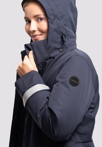 Chaqueta de marina resistente a la intemperie con capucha, tiras reflectantes plateadas en los puños, cierre delantero con cremallera y un parche circular con la marca en la manga.