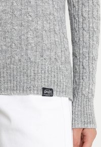 Gros plan sur une personne portant un pull gris en maille torsadée avec une étiquette noire près de l'ourlet et un pantalon blanc, la main détendue à son côté.