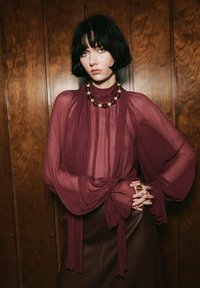 Bordeaux doorzichtige blouse met wijdvallende mouwen, geplooide details bij de taille en een hoge hals. Draagt met een bruine leren rok en een ketting van kralen.