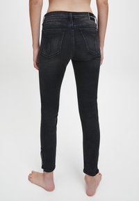 Jeans de mezclilla negra ajustados con una textura suave, cintura de altura media y bolsillos traseros curvados. Presentan detalles de cremallera en los dobladillos de los tobillos.