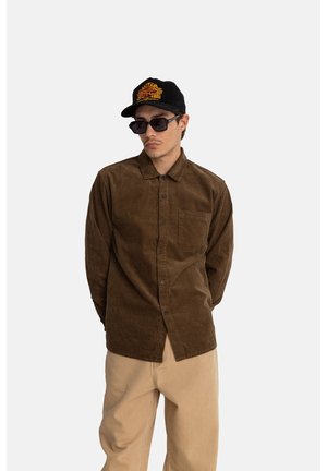 Brown corduroy shirt with long sleeves and a button-up front, paired with beige pants. Black cap features orange graphic design. Dark sunglasses.