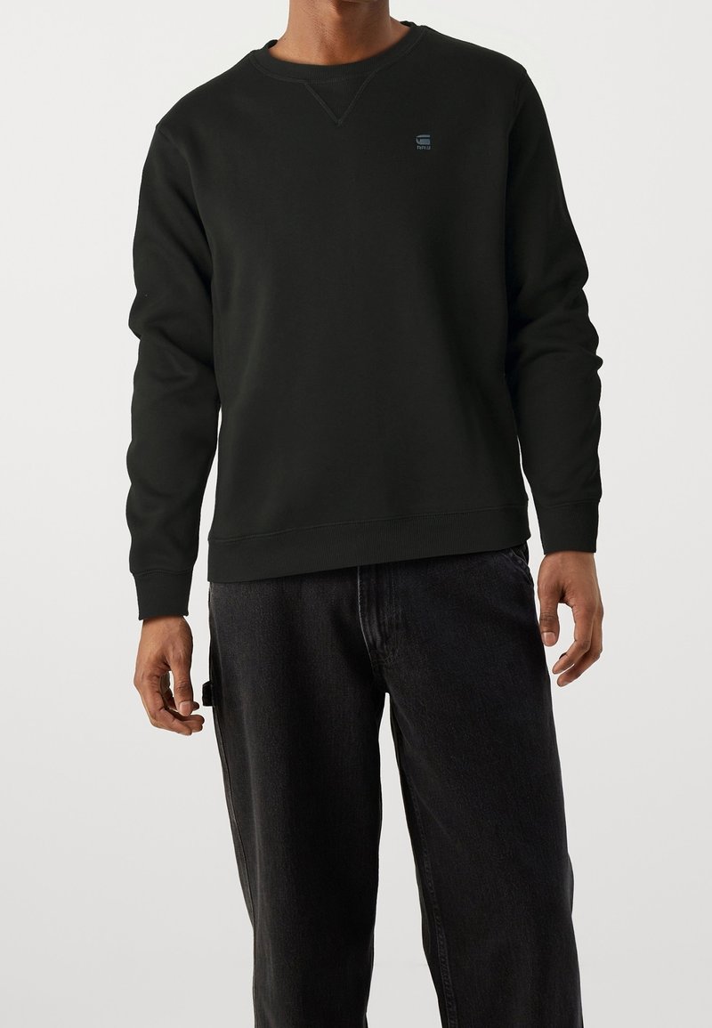 Sweat-shirt noir avec un col rond et des manches longues. Présente un petit logo sur la poitrine ainsi que des poignets et un ourlet côtelés. Porté avec un jean foncé.