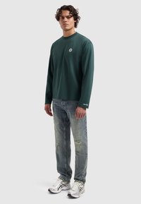 Langsleeve groen katoenen shirt met ronde hals en een klein logo op de borst, gedragen met lichtblauwe versleten jeans en zilveren sneakers.