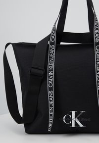 Czarna torba tote marki Calvin Klein Jeans z regulowanym paskiem na ramię i sznurkowymi rączkami z logo na białej powierzchni.