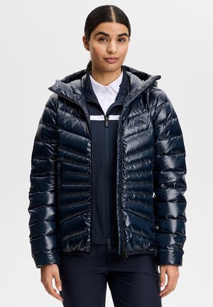 Kvinde med mørkt hår i en lav hestehale iført en skinnende marineblå quiltet jakke over en lynlåstrøje og en hvid skjorte med krave, stående foran en ensfarvet baggrund.