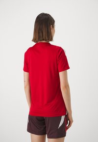 Camisa deportiva roja de manga corta, con cuello estándar y una textura suave. Combinada con pantalones cortos oscuros con detalles plateados.