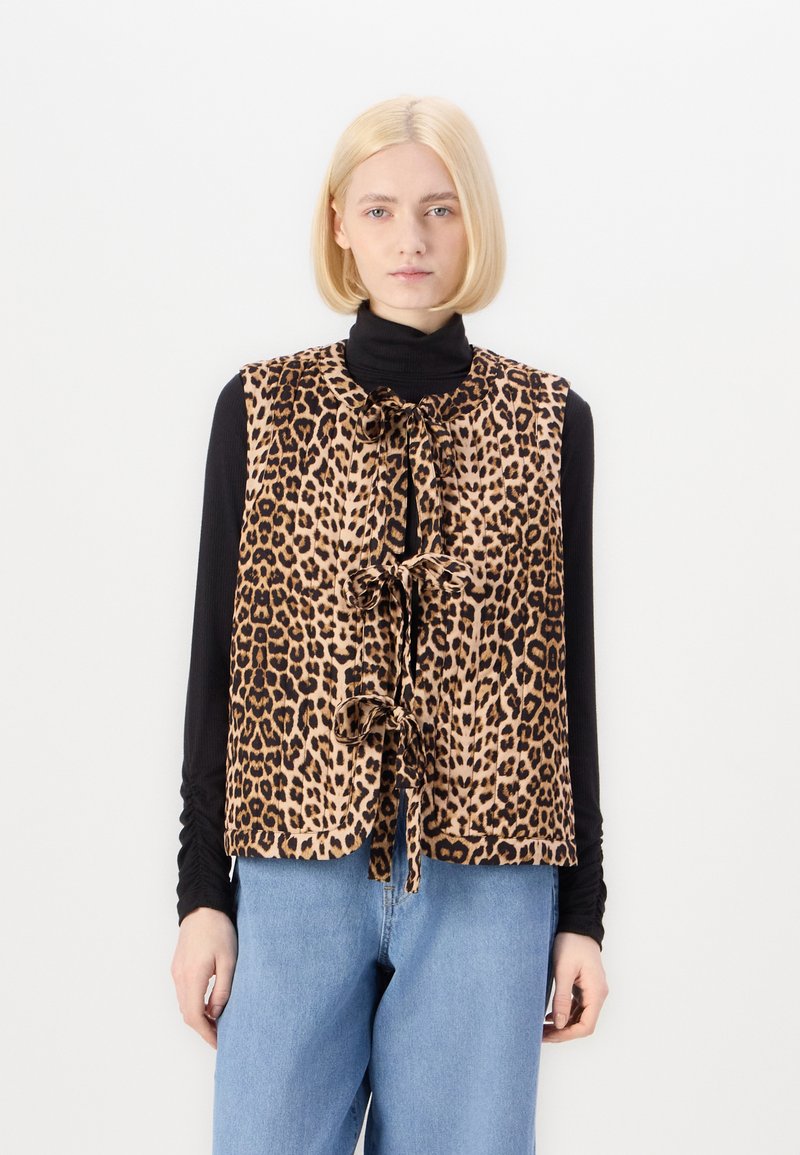 Γιλέκο με σχέδιο λεοπάρ, ελαφρύ ύφασμα, κουμπωτό μπροστινό μέρος με κορδόνια, χωρίς μανίκια, φορεμένο πάνω από μαύρη μπλούζα με μακριά μανίκια, σε στυλ με μπλε τζιν.
