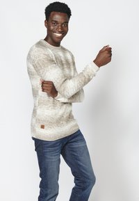 Lichtbeige textuurtrui met een subtiel breipatroon en ribbels aan de mouwen, gecombineerd met donkerblauwe jeans. Heeft een klein leren logo op de taille.