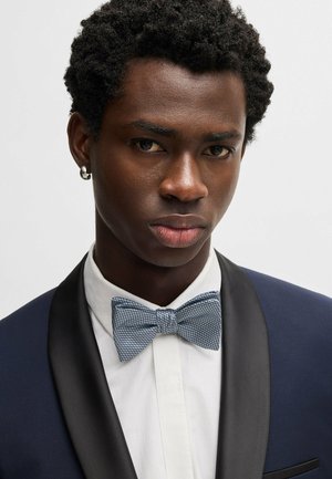 Abito blu navy con risvolto in raso, camicia bianca e papillon blu strutturato. Primo piano del viso con capelli corti e ricci e un orecchino.