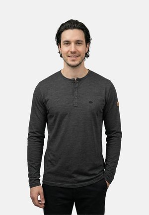 Mężczyzna o ciemnych włosach, ubrany w ciemnoszarą koszulę henley z długim rękawem i guzikami, stojący na tle jasnego, jednolitego tła, uśmiechający się.