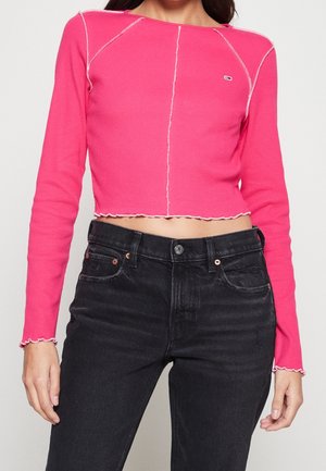 Femme portant un top court rose vif à manches longues avec coutures blanches apparentes et un jean noir taille haute, vue des épaules aux hanches.