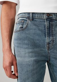 Jeans de mezclilla azul claro con un corte ajustado, que cuentan con un diseño clásico de cinco bolsillos, costuras en marrón contrastante y un cierre de botón metálico.