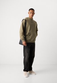 Jonge man staat rechtop in een olijfgroene hoodie, zwarte jeans, beige sneakers en draagt een zwarte schoudertas, tegen een effen witte achtergrond.