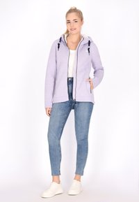 Felpa con zip di colore viola chiaro, dotata di cordini a contrasto, tasche laterali e polsini a coste, indossata sopra una maglietta bianca e abbinata a jeans blu.