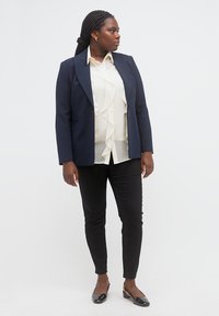 Γυναίκα στέκεται κοιτώντας δεξιά, φορώντας μπλέ σκούρο σακάκι, λευκό μπλουζάκι με βολάν, μαύρο παντελόνι και μαύρα παπούτσια με χαμηλό τακούνι, πάνω σε ένα απλό φόντο.