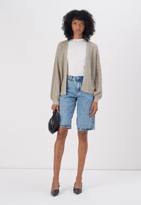 Lichtbruine, gebreide cardigan met gevallen schouders, gecombineerd met hoge spijkerbroek shorts. Model houdt een zwarte handtas vast en draagt hakken.