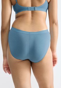 Vrouw in blauwe naadloze onderbroek en bijpassende bh, van achteren gezien tegen een witte achtergrond.